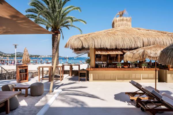 Außenbereich der Zel Mallorca Strandbar mit Strohdach, Tischen, Stühlen und Sonnenliegen.
