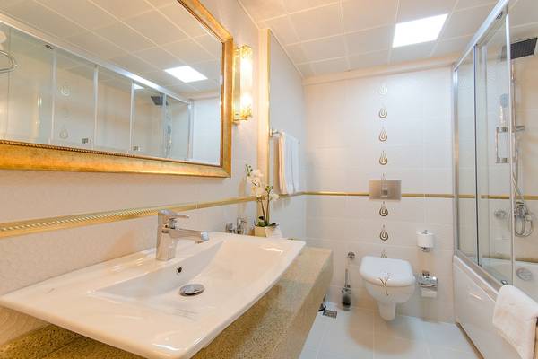 Badezimmer im Arden City Hotel mit weißen Fliesen, goldenen Akzenten, Waschbecken und Spiegel.