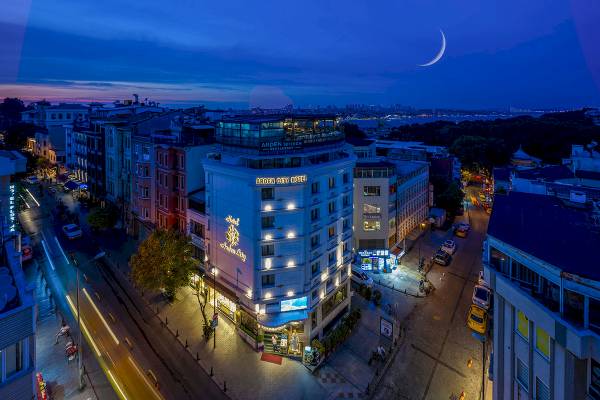 Abendliche Stadtansicht des Arden City Hotel mit beleuchteten Gebäuden und einer Halbmond im Himmel.