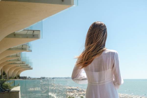 Rückansicht einer Frau in einem weißen Kleid, das auf einem Balkon mit Meerblick steht.