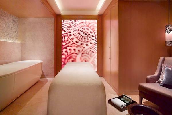 Behandlungsraum im Wellnesscenter des Grand Hyatt Dubai mit einem Behandlungstisch, einem Liegebett, einem Stuhl, einem Tisch mit Pflegeprodukten und einer Wand mit einem Mandala-Design.