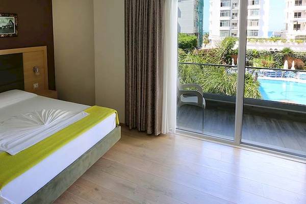 Dyrrah Hotel Zimmer mit Balkon und Poolblick zeigt ein gemütliches Bett, zwei weiße Kissen, eine grüne Decke, eine Glastür und einen Swimmingpool.