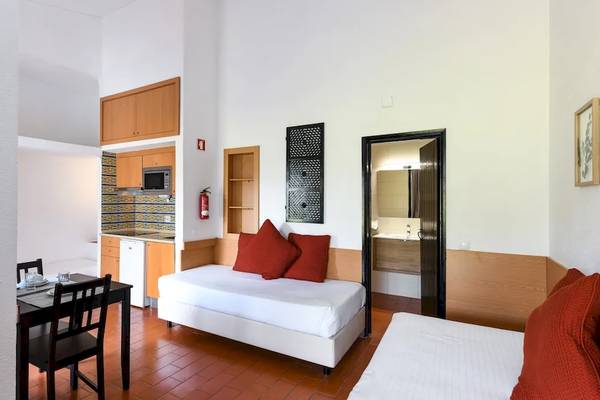Innenansicht eines Hotelzimmers im Pedras da Rainha Resort mit einem Bett, Tisch, Stühlen und Küche.