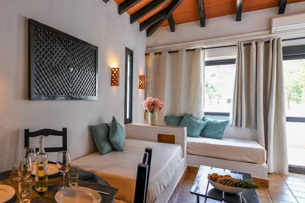 Innenansicht eines Zimmers im Pedras da Rainha Resort mit Esstisch, Sesseln, Bett und Fenstern.