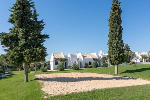 Außenbereich des Pedras da Rainha Resorts mit einem Volleyballnetz, umgeben von grünem Rasen und hohen Bäumen.