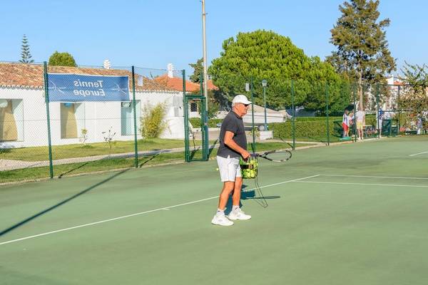 Tennisplatz im Pedras da Rainha Resort mit einem Mann, der Tennisschläger und Ballkorb hält.