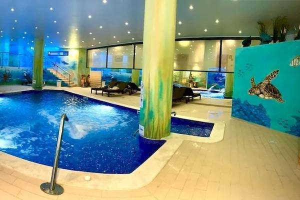 Indoor Swimming Pool im Baia Grande Hotel mit blauem Wasser, bunten Wandmalereien und Sonnenliegen.