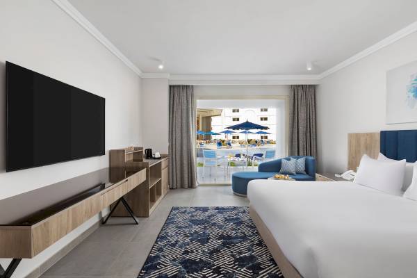 Innenansicht eines Hotelzimmers im Pickalbatros Blu Spa Resort mit Fernseher, Schrank, Teppich und Blick auf den Pool.