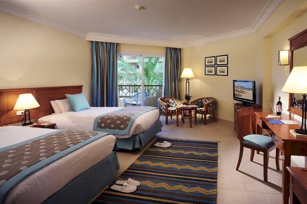Doppelzimmer im Amwaj Beach Club mit zwei Betten, Fernseher und Balkon mit Aussicht auf Palmen und Gebäude.