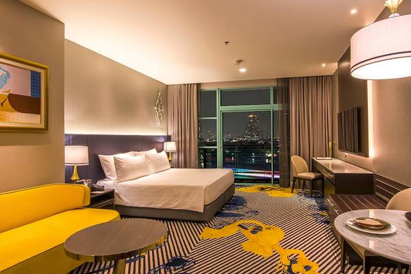 Innenansicht eines Zimmers im Chatrium Hotel Riverside Bangkok mit Bett, Couch, Schreibtisch und Aussicht auf die Stadt.