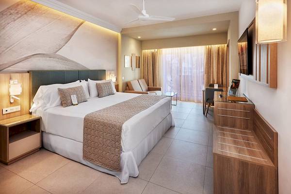 Schlafzimmer im RIU Palace Macao mit großem Bett, Kissen, Decken und Holzmöbeln.