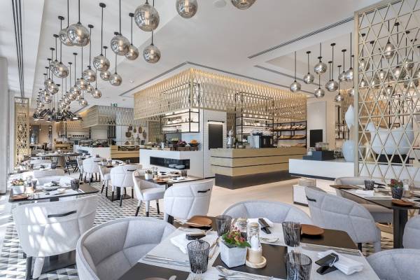 Innenansicht des Al Bandar Rotana Restaurants mit weißen Stühlen und Tischen, goldenen Leuchten und einem langen Tresen.