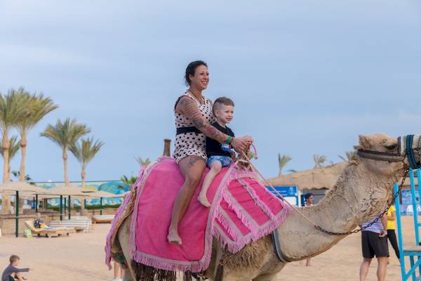 Eine Frau und ein Kind reiten auf einem Kamel am Strand, umgeben von Palmen und weiteren Besuchern.
