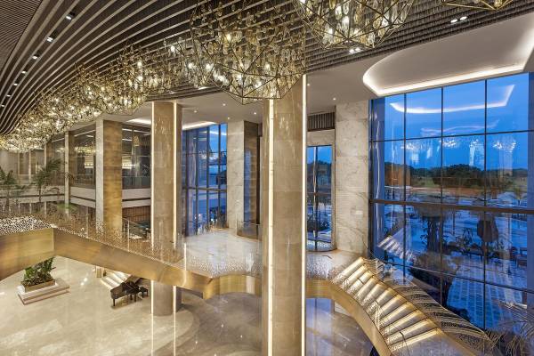 Empfangsbereich des Cullinan Belek Hotels mit goldenen Kronleuchtern und großen Fenstern.