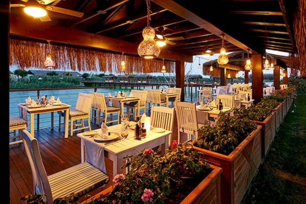 Außenbereich des Restaurants im Gloria Serenity Resort mit Blick auf das Wasser und grüne Pflanzen.