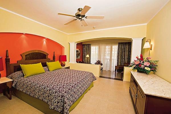 Schlafzimmer im Iberostar Selection Rose Hall Suites Hotel mit gelben und orangefarbenen Wänden, einem großen Bett, grünen Kissen und einem Deckenventilator.