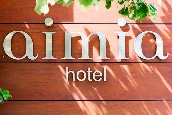 Schild des Aimia Hotels an einer braunen Holzwand mit Schatten und grünen Blättern