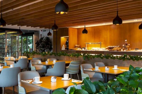 Frühstücksbereich des Aimia Hotels mit Tischen, Stühlen und Pflanzendekor.