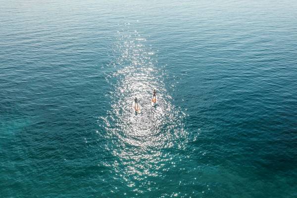 Zwei Personen auf Surfbrettern im kristallklaren Wasser vor einer blauen Himmelslandschaft.