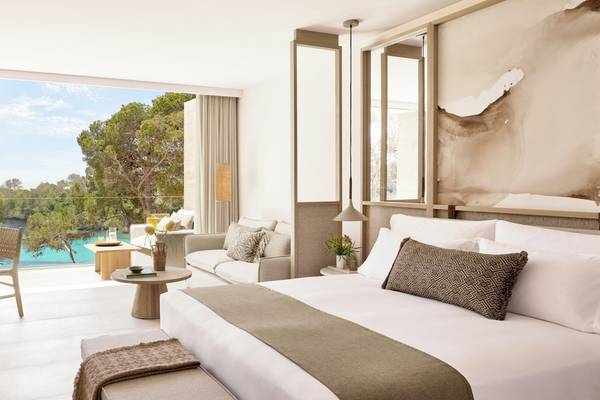 Ein Hotelzimmer im Ikos Porto Petro mit großem Fenster, Blick auf einen Pool, Möbeln und Pflanzen.