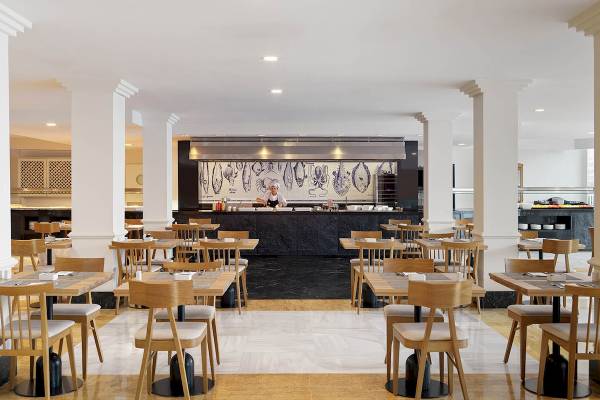 Innenansicht des Restaurants des H10 Blue Mar Boutique Hotels mit leeren Tischen und Stühlen und einem Koch hinter der Theke.