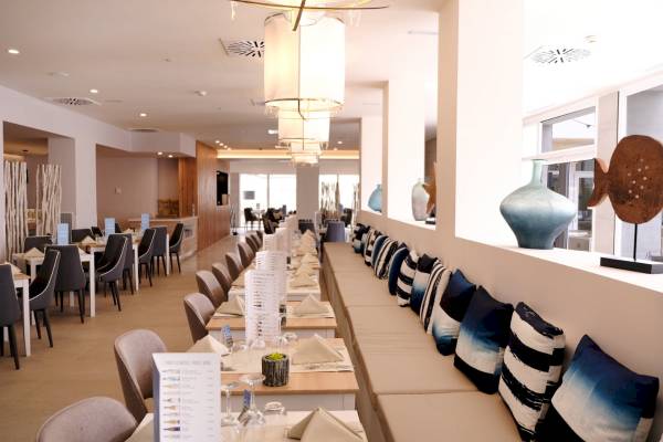 Innenansicht des Restaurants im Nautic Hotel & Spa mit langen Tischen, Stühlen, blau-weiß gestreiften Kissen und einem großen Fenster.