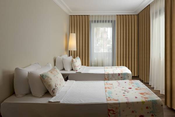 Zwei Betten im Zimmer des Seven Seas Hotel Life, beide mit bunten Decken und weißen Kissen, neben einem Fenster mit Jalousien und einem Lampenschirm.