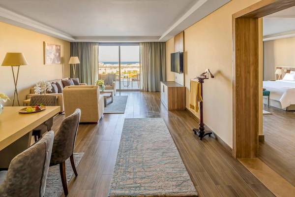 Innenansicht eines eleganten Hotelzimmers der Rixos Premium Magawish Suites & Villas mit Wohnzimmer, Essbereich und Schlafzimmer.