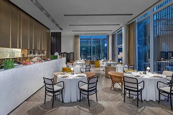 Innenbereich des Restaurants im Oberoi Beach Resort, Al Zorah, mit runden Tischen und Stühlen, großen Fenstern und Pflanzen.