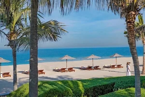 Strand des Oberoi Beach Resorts Al Zorah mit Liegestühlen, Sonnenschirmen und einer Person in einem weißen Kleid.