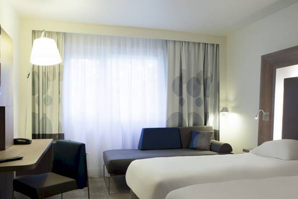 Innenansicht eines Hotelzimmers im Novotel London Canary Wharf mit Schreibtisch, Stuhl, Sofa und zwei Betten.