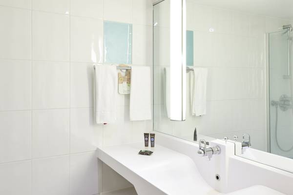 Badezimmer im Novotel London West Hotel mit weißen Fliesen, Spiegel und Handtüchern.