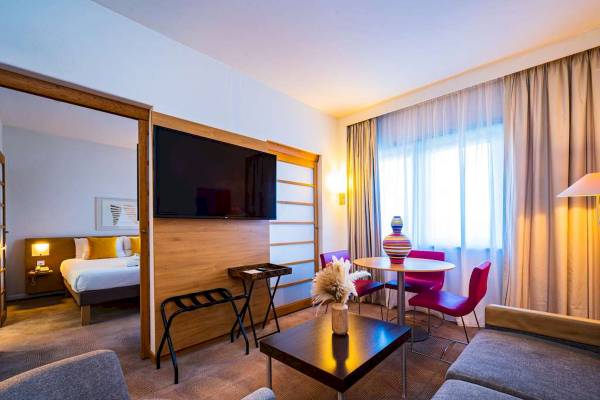 Innenansicht der Suite im Novotel London West mit Flachbildfernseher, Bett, Tisch, Stühlen und Wohnzimmermöbeln.