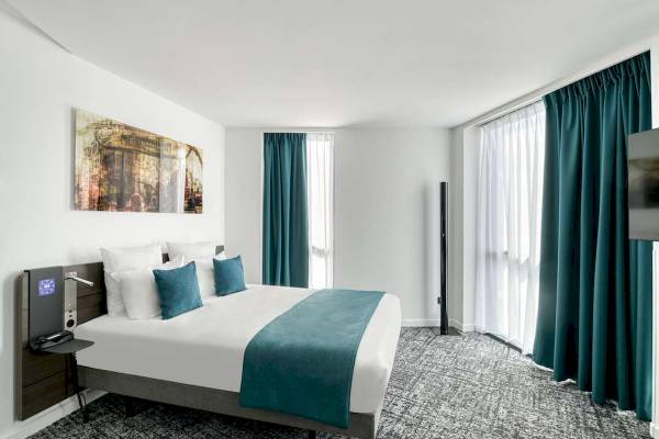 Innenansicht eines Hotelzimmers im Novotel Paris Centre Bercy mit Bett, Kissen, Gemälde und grünen Vorhängen.