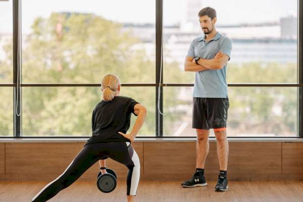 Eine Frau macht eine Übung mit einer Hantel in einem Fitnessstudio, während ein Mann zuschaut.