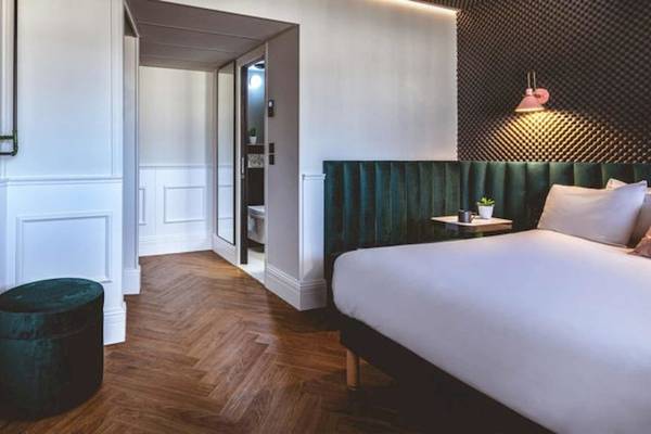 Innenansicht eines Hotelzimmers im The ReMIX Hotel mit Holzboden, weißen Wänden, einem Bett und einem grünen Hocker.