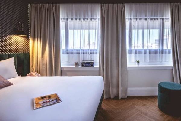 Schlafzimmer im ReMIX Hotel mit großem Bett, weißem Laken, zwei Fenstern, einem grünen Hocker und einem Fernseher an der Wand.