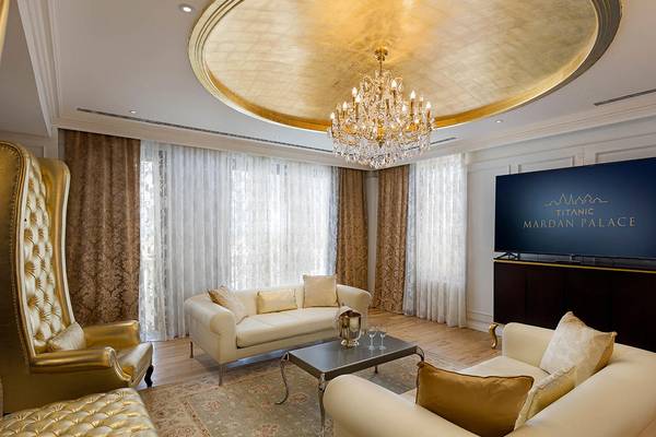 Wohnzimmer des Titanic Mardan Palace mit goldener Decke, Kristallkronleuchter, weißen Sofas, Couchtisch, Fernseher und goldenen Stühlen.