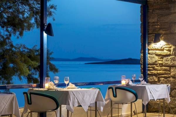 Außenbereich des Mykonos Theoxenia Boutique Hotels mit Tischen und Stühlen, die auf eine Aussicht auf das Meer und Inseln ausgerichtet sind.