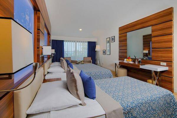 Zwei Doppelbetten in einem Zimmer im Limak Atlantis Deluxe Hotel & Resort mit blauen Vorhängen und Holzmöbeln.
