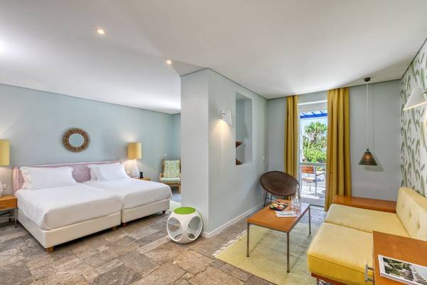 Schlafzimmer im Mykonos Theoxenia Boutique Hotel mit zwei Betten, einem Couchtisch, einem Stuhl und einem Fenster mit gelben Vorhängen.