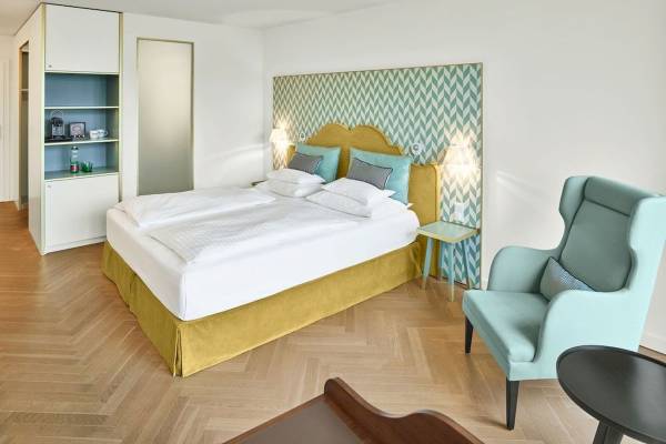 Schlafzimmer im Maxx by Steigenberger Wien mit einem Bett, Nachttisch, Sessel und einer blauen Wandgestaltung.