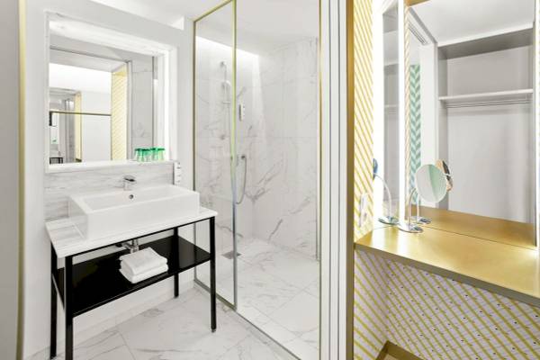 Das elegante Badezimmer des MAXX by Steigenberger Vienna mit weißen Marmorfliesen, goldenen Akzenten und einer modernen Dusche.