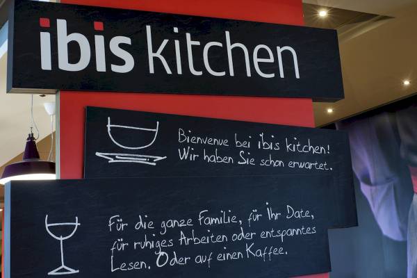 Schwarze Tafeln an einem Stand der Ibis Kitchen mit Willkommensgruß auf Deutsch.