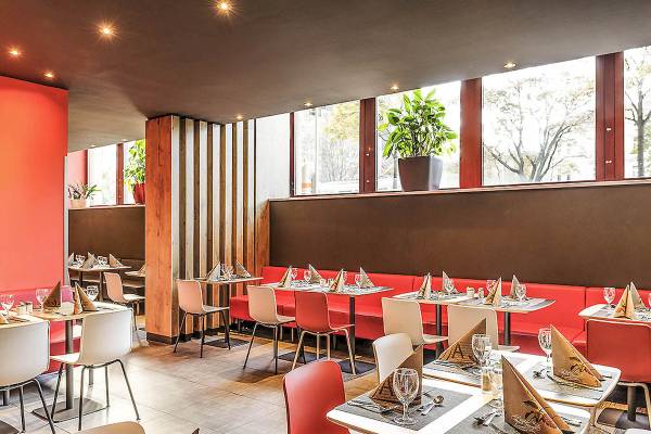 Innenansicht des Restaurants im Ibis Wien Mariahilf mit roten Bänken und Tischen, Fenstern und Pflanzen.
