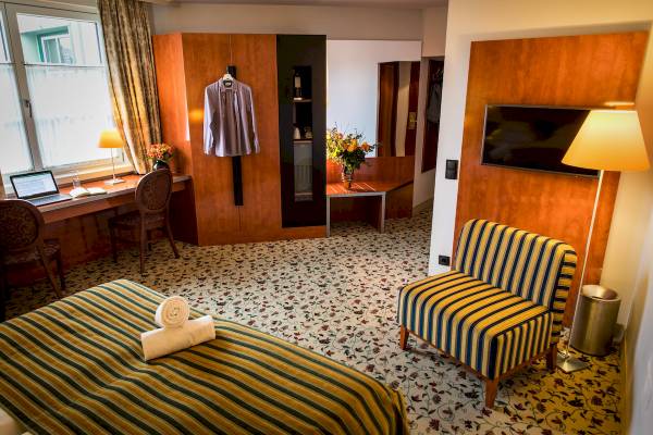 Innenansicht eines Zimmers im Mercure Grand Hotel Biedermeier mit Schreibtisch, Stühlen, Bett, Fernseher und Dekor.