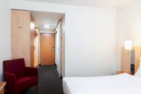 Innenansicht eines Hotelzimmers im IntercityHotel Wien mit einem Bett, einem roten Stuhl und einer Lampe.