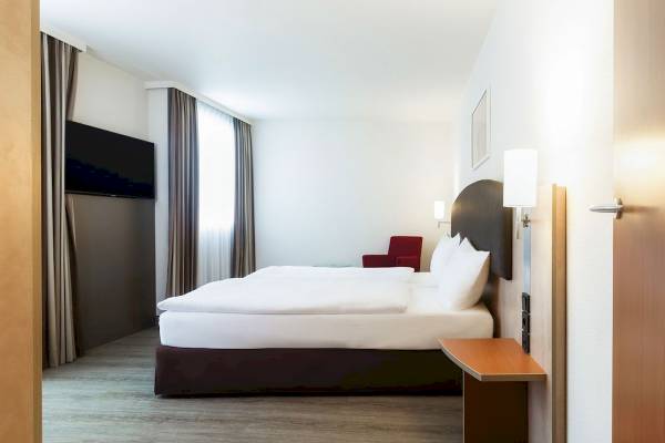 Doppelbettzimmer im IntercityHotel Wien mit Fernseher, rotem Stuhl und Lampen.
