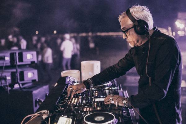 Ein älterer DJ mit weißer Frisur und schwarzer Kleidung spielt Musik in einer Nachtclub-Umgebung.
