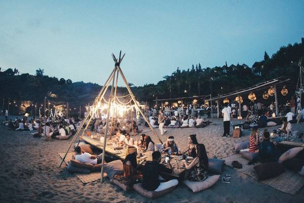 Eine Gruppe von Menschen, die auf Kissen am Strand sitzen und essen, mit einem großen Tipi und Lichterketten im Hintergrund.